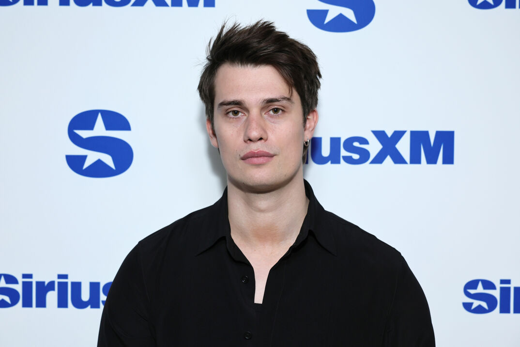 Nicholas Galitzine poses in front of a step-and-repeat. He has spiked dark brown hair and wears a hoop earring in his left ear. He's pictured wearing a black collared shirt over a black low cut tank top.