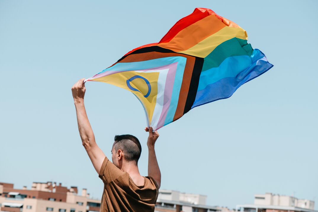 A person holding a Progress Pride Flag aloft against a clear sky, with the flag featuring the traditional six-striped rainbow design plus additional colored chevrons representing marginalized LGBTQ+ communities, including black and brown stripes for marginalized POC communities, and the colors pink, light blue, and white, symbolizing trans and non-binary individuals.