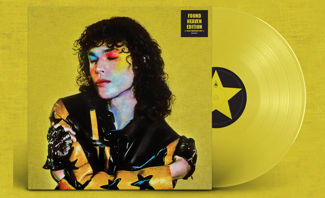 The vinyl album for Conan Gray's 'Found Heaven,' featuring the black haired singer closing his eyes with rainbow makeup around his eyes in a leather jacket hugging himself. He's pictured in front of a yellow wall and a yellow vinyl disc peeks out of the sleeve.