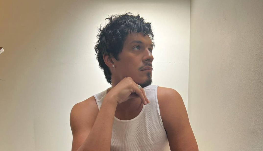 Omar Apollo sits in a white tank top looking off. He has an earring in his right ear, messy black hair, and rests his hand on his chin