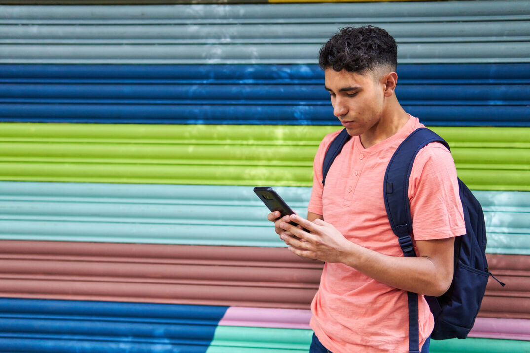 A young Latino man with his backpack answers his mobile phone walking in the streets of Mexico City, wearing blue pants and a melon shirt.