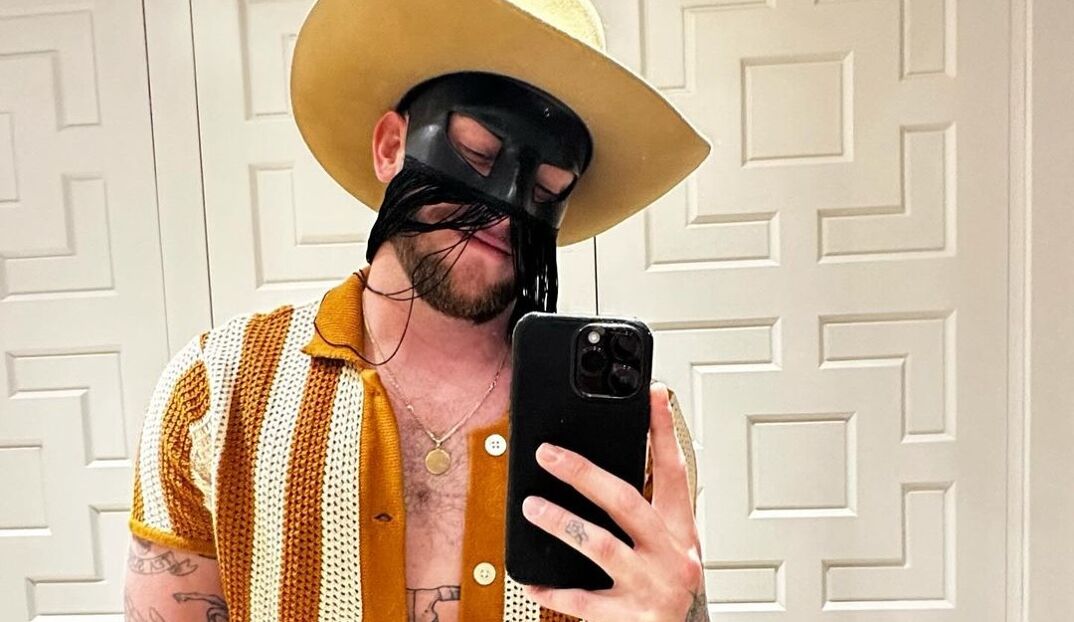 Orville Peck takes a selfie in a bathroom mirror on his iPhone. He wears a yellow cowboy hat, a black mask obscuring the top half of his hat, and a crocheted orange and white collared shirt, unbuttoned to show off the top of his muscular chest.