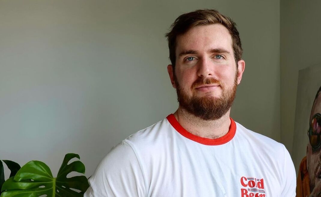 The content creator known as Girthmaster stands smiling in his living room. he has blue eyes, reddish brown hair and a full beard, and smiles softly. He wears a white tee shirt with a red collar that reads "Cold Beer."