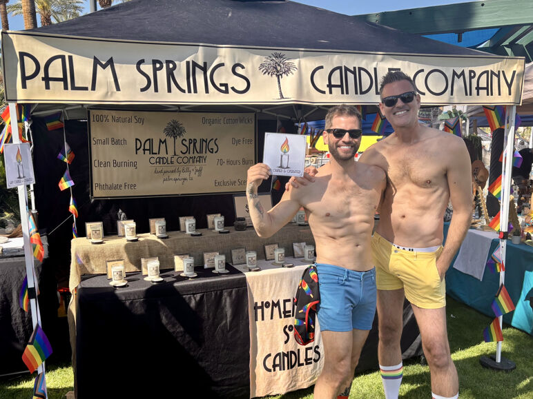 Billy and Jeff, the married couple behind Palm Springs Candle Company, pose shirtless in colorful shorts and sunglasses in front of their outdoor market booth. The booth displays their handmade soy candles with a sign listing features like “100% Natural Soy,” “Small Batch,” and “Phthalate Free.” Pride flags and rainbow details surround the setup on a sunny day.