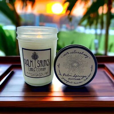A Palm Springs Candle Company jar candle and its matching lid labeled “Palm Springs Sunrise” are displayed on a wooden tray at sunset. The background features palm trees, soft orange sunlight, and a blurred view of water. The candle is labeled “hand poured by Billy & Jeff, Palm Springs, CA.”