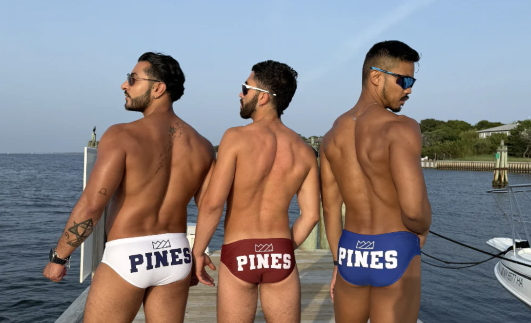 Three shirtless men pose on the dock at Fire Island Pines. They are wearing the same pair of swim briefs in different colors, which reads "PINES" on the backside.
