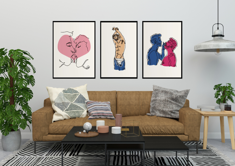 A modern living room with a brown suede couch, patterned pillows, and a black coffee table, featuring a gallery wall of three framed queer art prints by JustMikeysArt. The prints show: a minimalist pink line drawing of two people kissing, a shirtless figure stretching in blue jeans and white underwear, and a colorful portrait of two masculine figures facing each other in blue and pink tones. A large leafy plant and industrial-style light fixture complete the space.