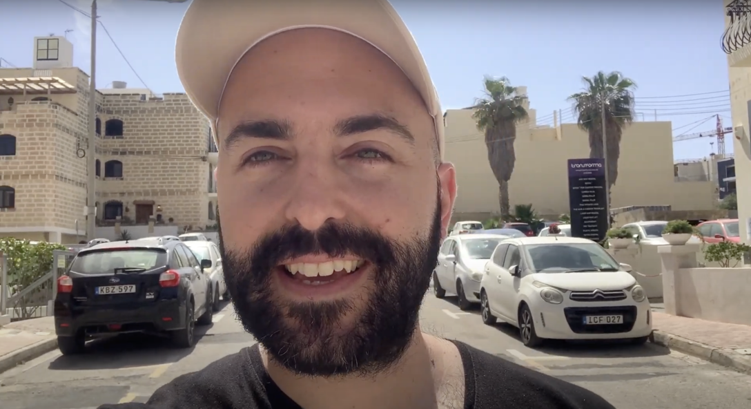 Matthew Grech walks around outside in Malta talking into a video camera wearing a baseball hat.