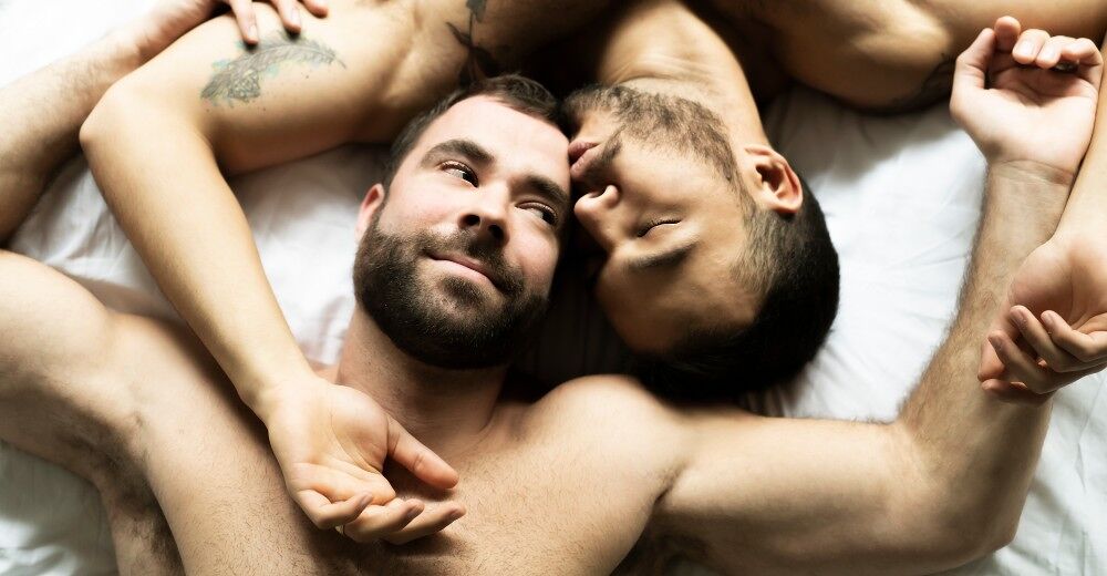 Two gay men lie entangled in bed