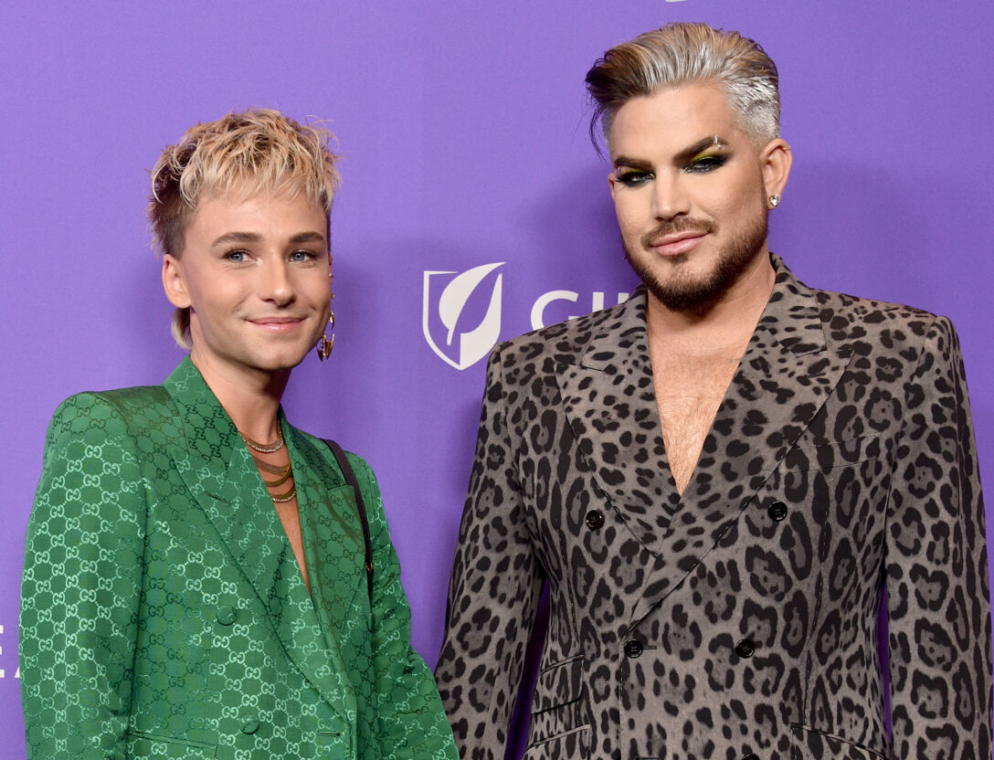 Oliver Gliese in a green suit jacket stands next to Adam Lambert in a leopard-print low-cut suit jacket on the red carpet.