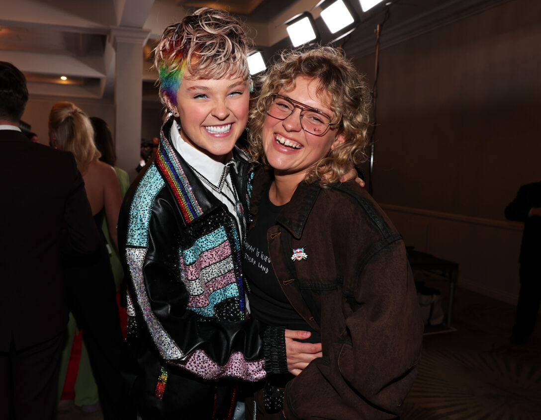JoJo Siwa, with a messy blonde and rainbow-colored pixie cut, hugs her then-partner Kath Ebbs backstage at an event.