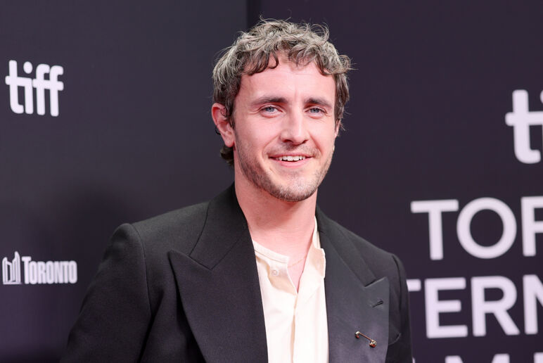 Paul Mescal smiles softly in a black suit and white dress shirt on the red carpet.