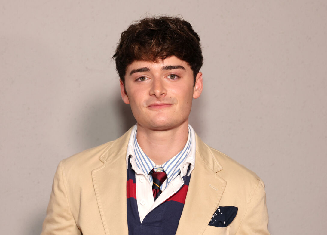 Noah Schnapp smiles softly in front of a tan background in a button down t-shirt, tie, and tan blazer.
