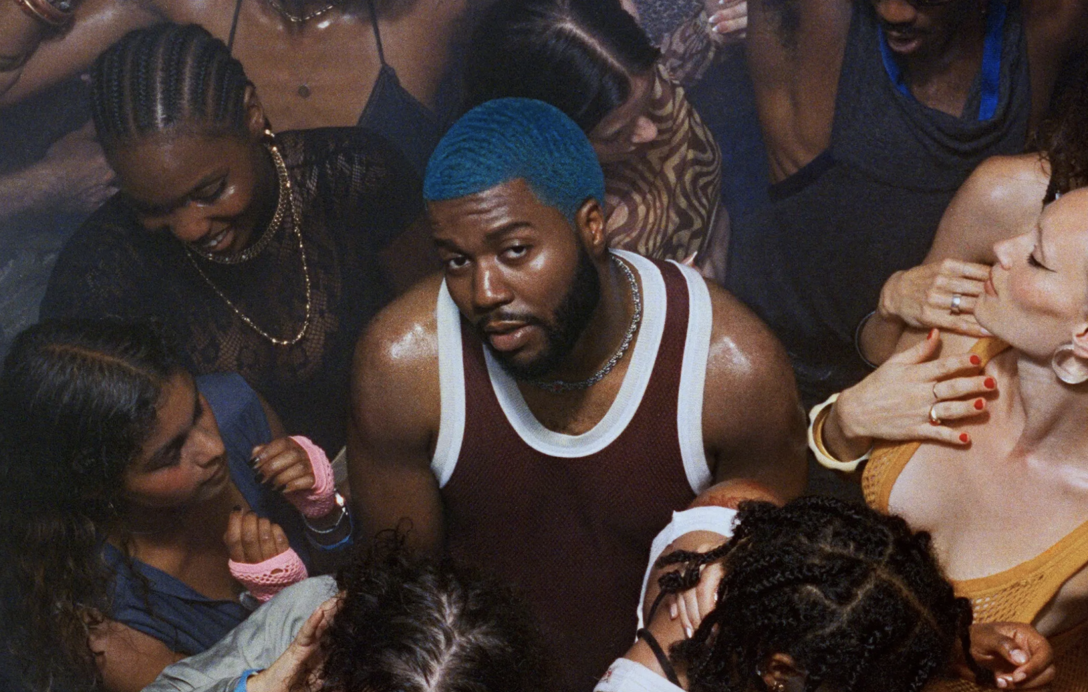 Khalid stands in the middle of a sweaty dance floor with blue hair in a red tank top, looking up mischievously.