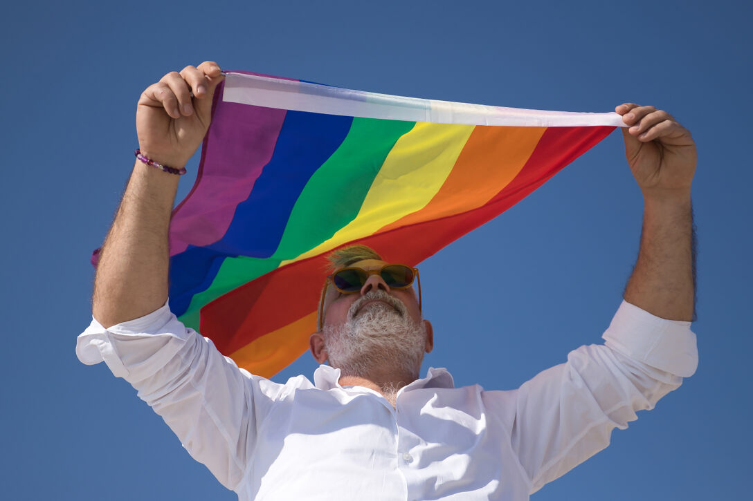 A gay man with a white beard stands solemnly under a rainbow flag in front of a blue sky.